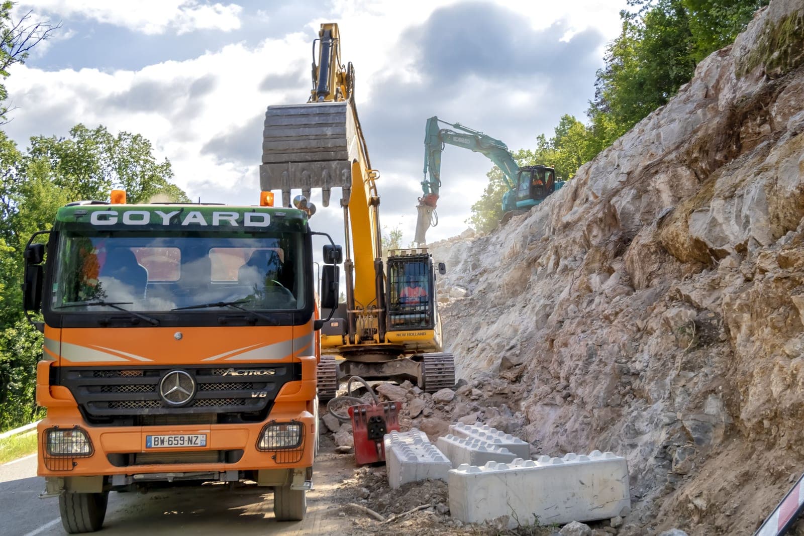 Camion-benne Goyard et pelles hydrauliques travaillant au confortement d'un talus rocheux le long d'une route.