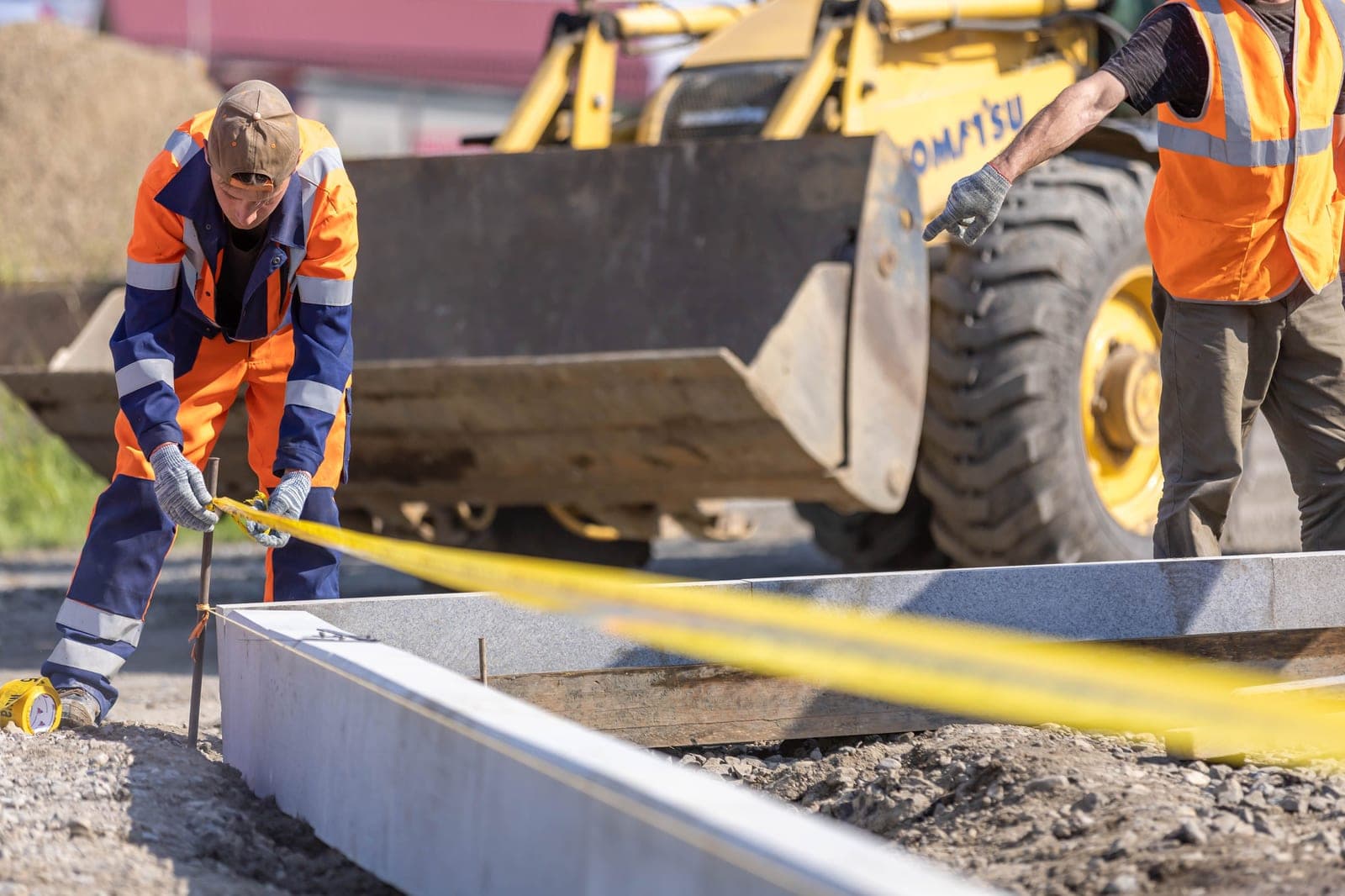 Deux ouvriers de travaux publics implantent une bordure de voirie au cordeau devant une chargeuse Komatsu.